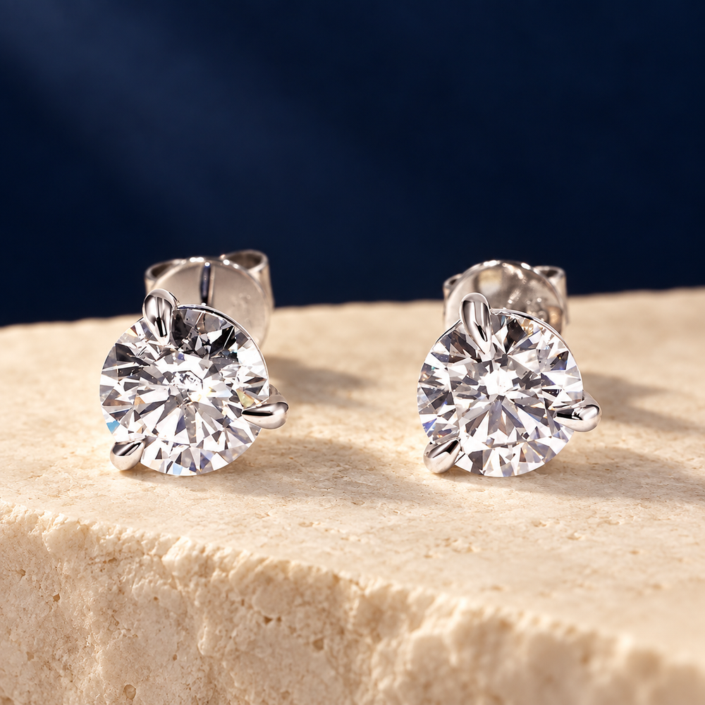 Three-claw diamond stud earrings styled on pale stone against midnight blue.